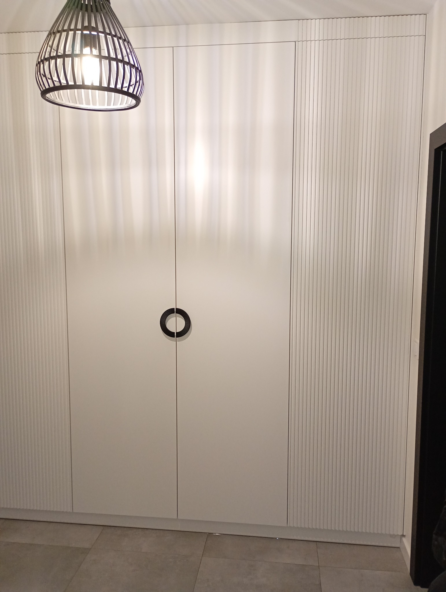 Biała szafa wnękowa z ryflowanymi bokami i okrągłymi uchwytami, oświetlona lampą z ażurowym kloszem. Widoczna szara podłoga i fragment czarnej framugi drzwi.