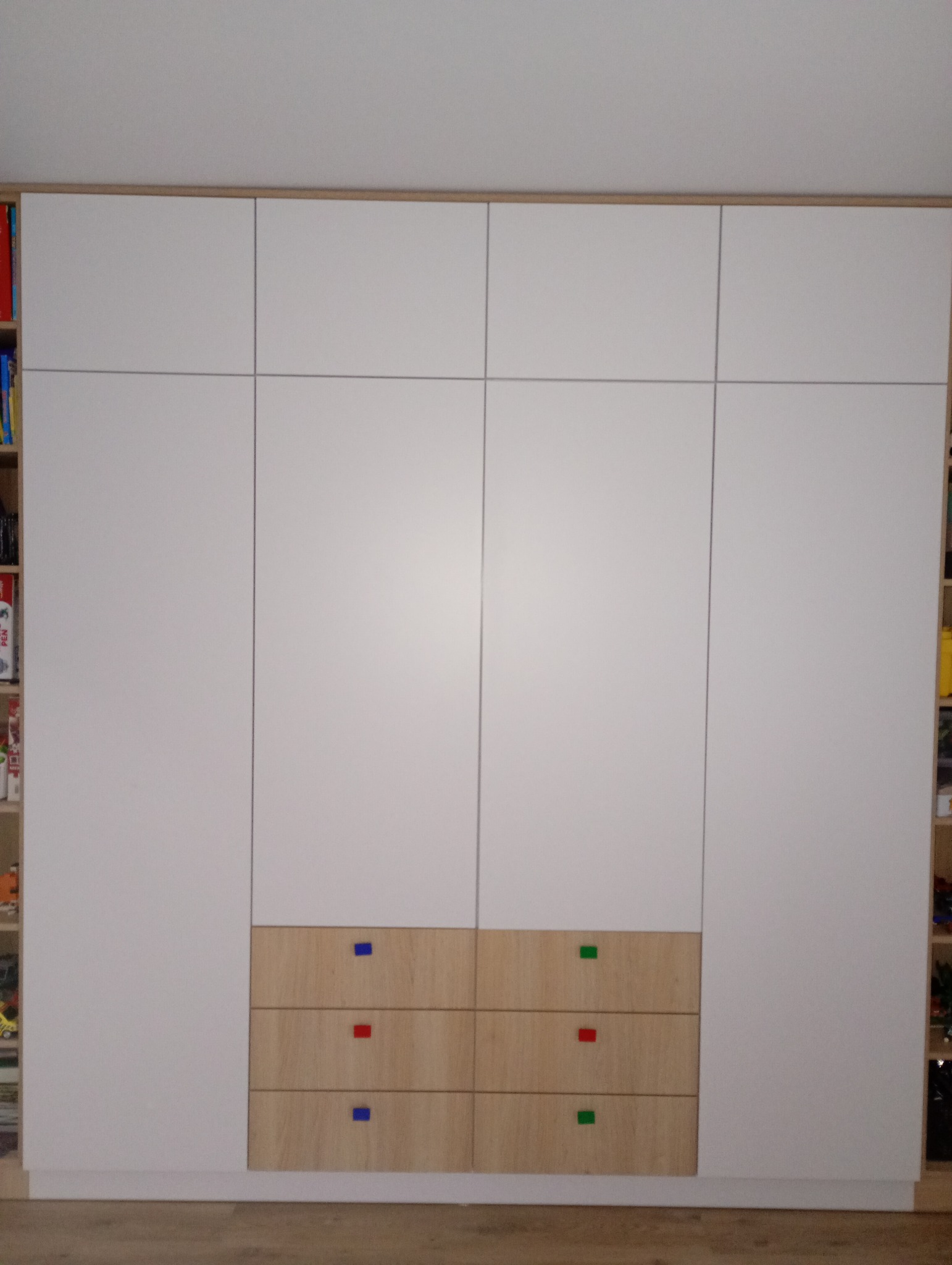 Biała szafa wnękowa z drewnianymi szufladami i kolorowymi uchwytami (niebieski, czerwony, zielony). Po bokach regały z książkami i zabawkami.