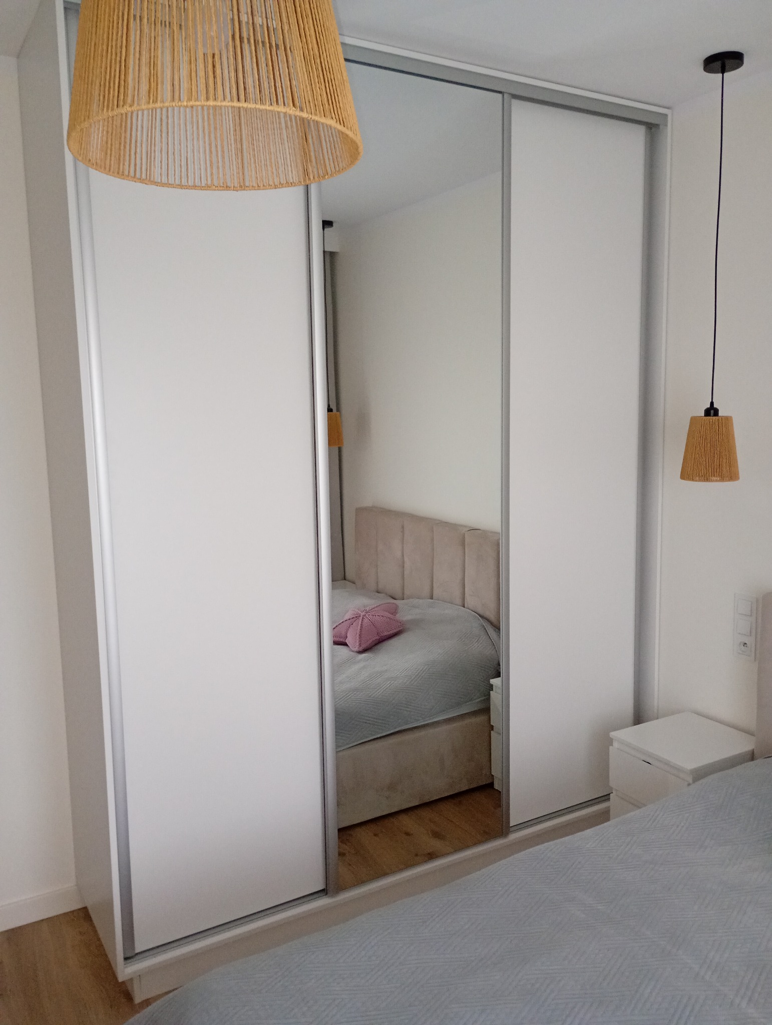 Biała szafa przesuwna z lustrem w sypialni, odbijająca łóżko z różową poduszką. W tle lampy z abażurami z naturalnego materiału. Minimalistyczny design.