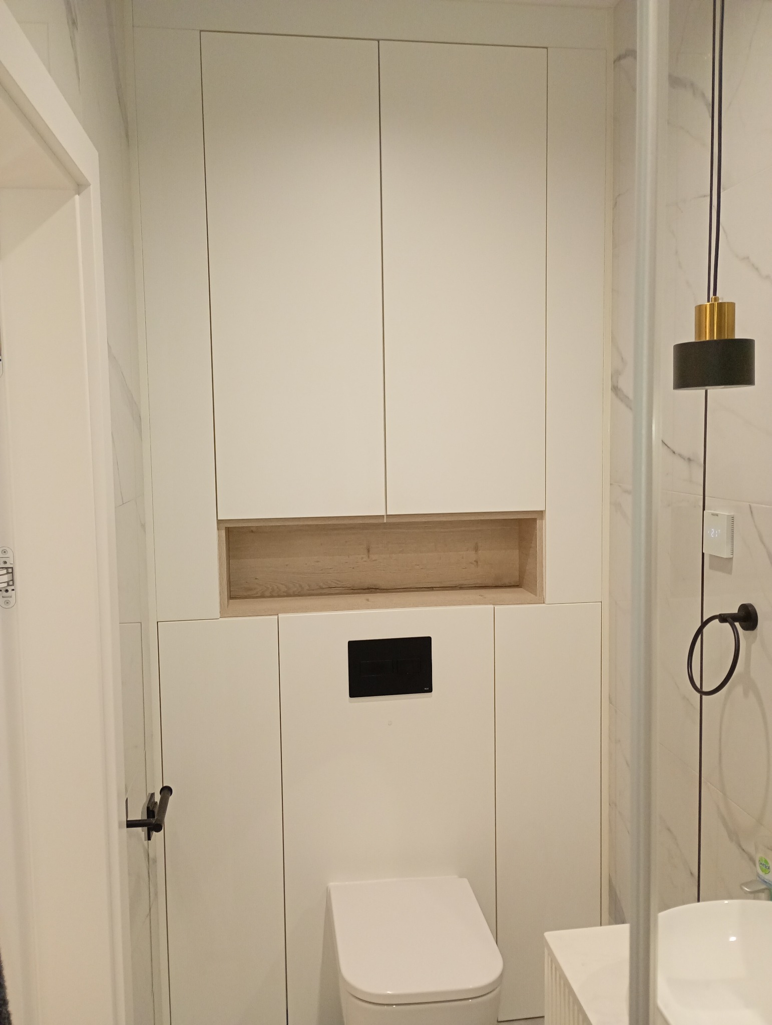 Nowoczesna, minimalistyczna łazienka z zabudową WC i szafkami w kolorze białym, wykończona marmurowymi płytkami. Czarny panel sterujący i oświetlenie.
