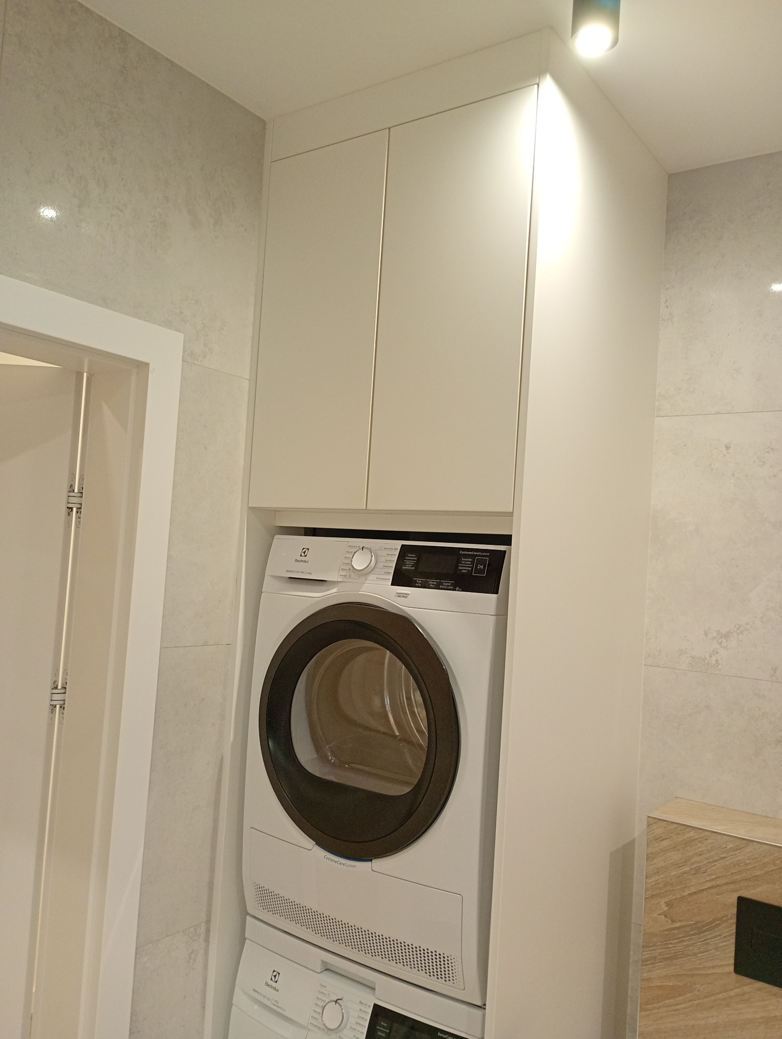 Biała pralka i suszarka Electrolux w zabudowie meblowej z szafkami w łazience wykończonej płytkami imitującymi beton. Oświetlenie punktowe.