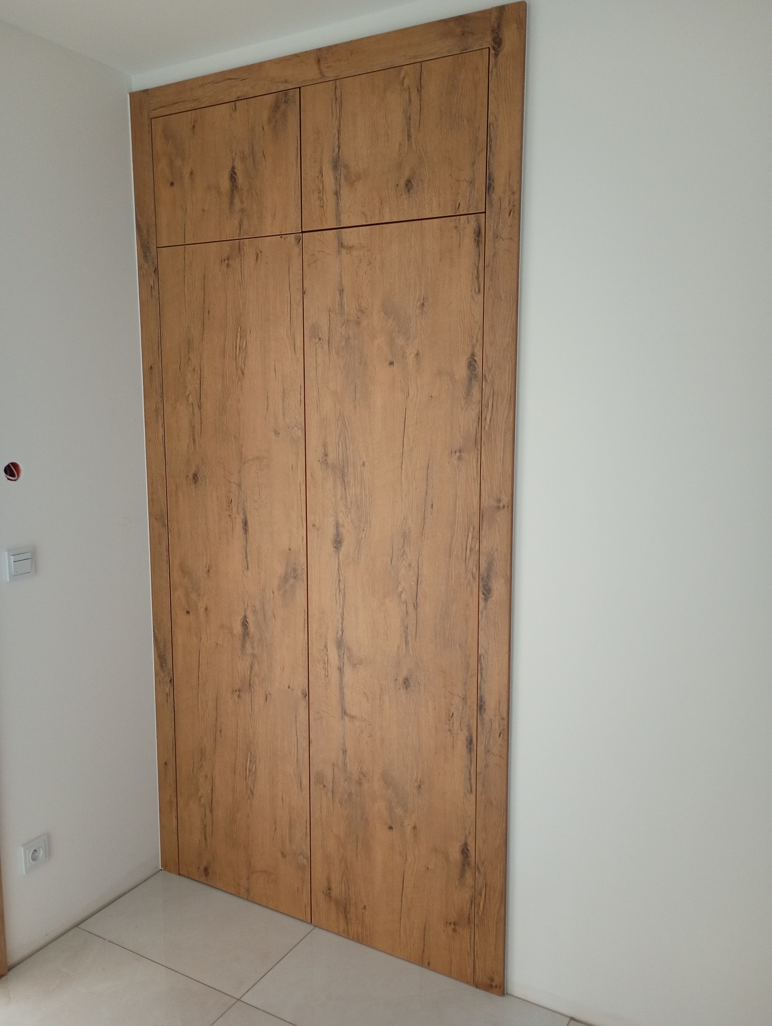 Wbudowana szafa z drewnianymi frontami, zamykana na całej wysokości ściany, wkomponowana w białe wnętrze. Widoczna faktura drewna i minimalistyczny design.