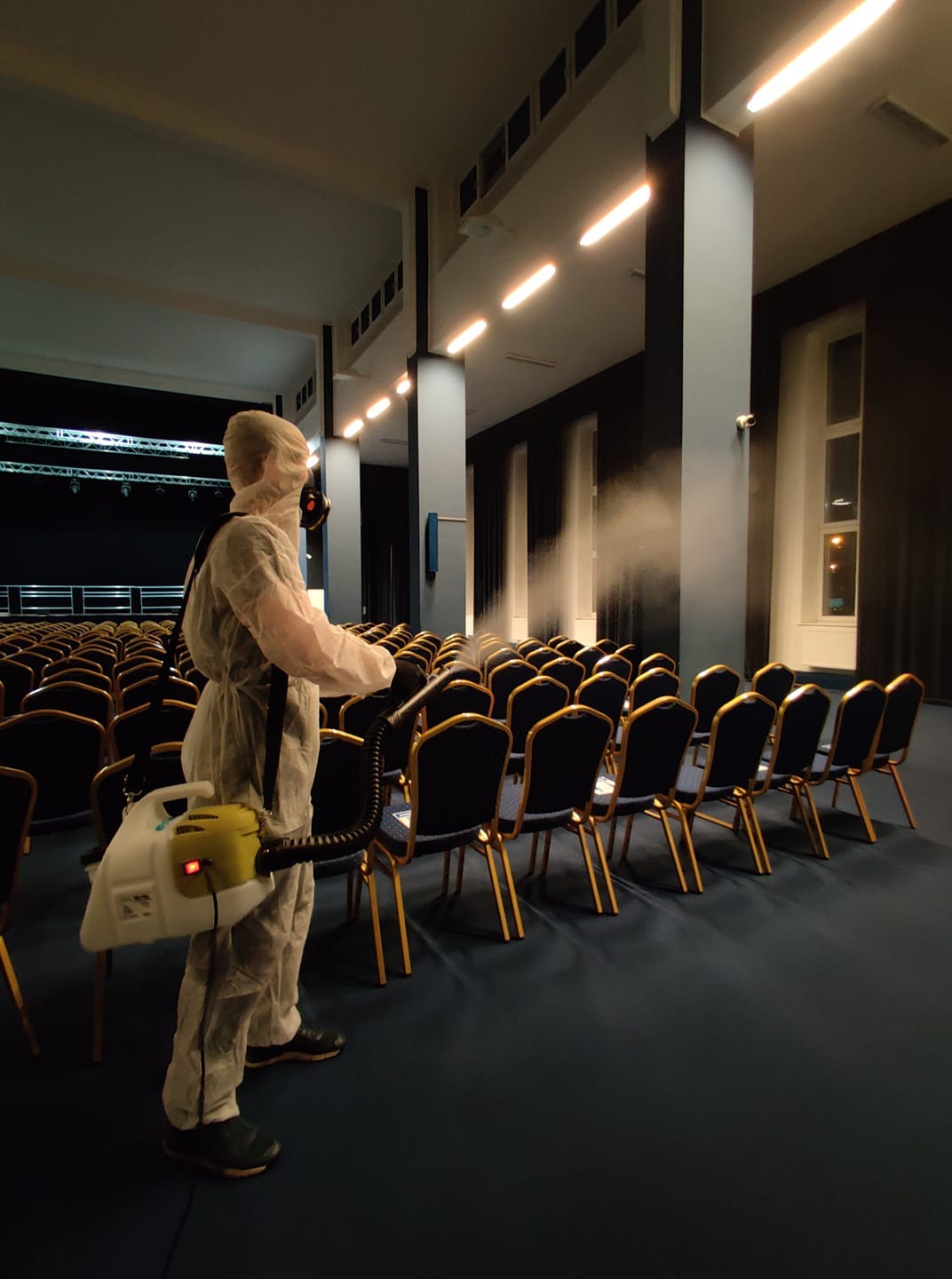 Osoba w kombinezonie ochronnym dezynfekuje krzesła w dużej sali za pomocą zamgławiacza, widoczne rzędy krzeseł, kolumny i oświetlenie sufitowe.