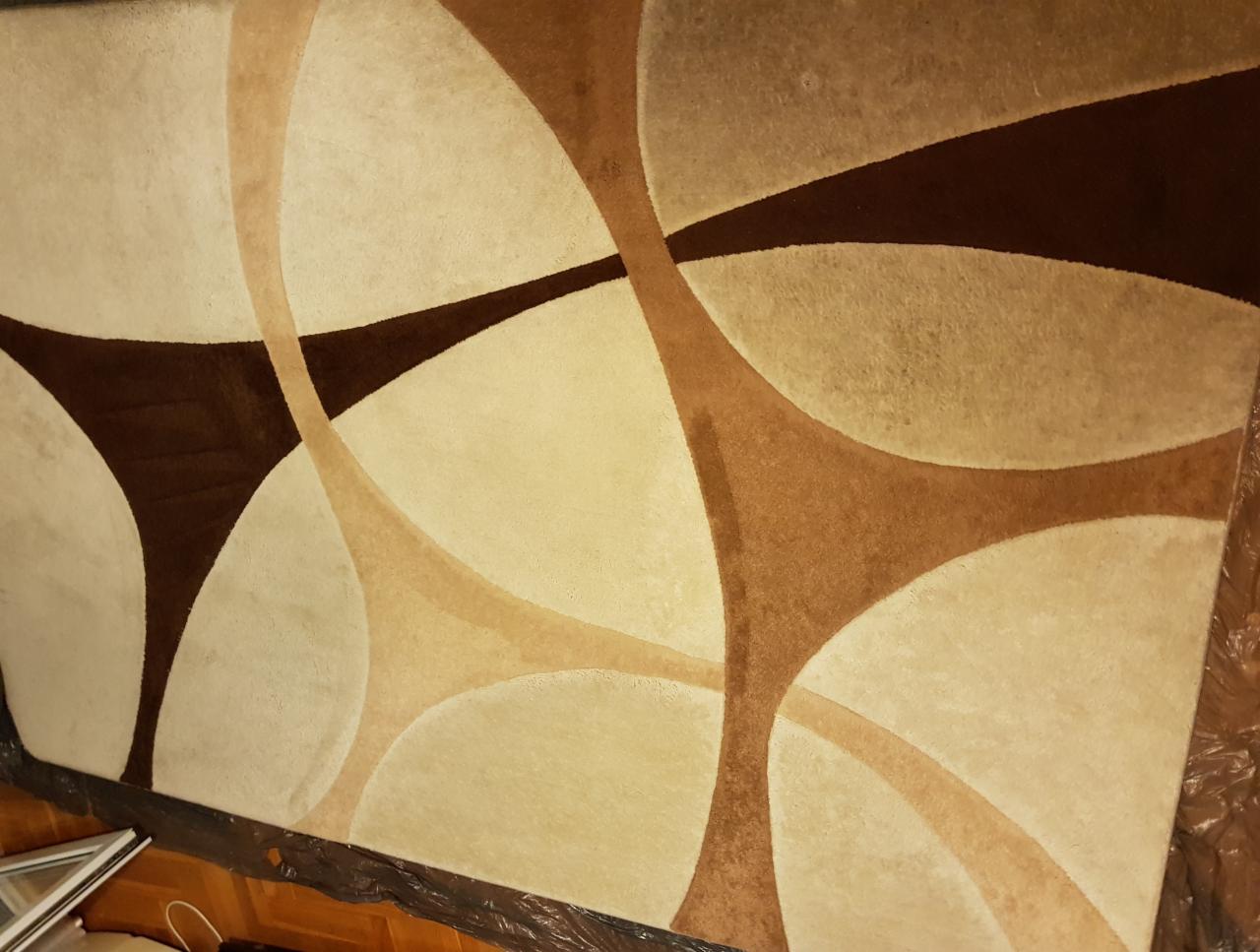 Wzór dywanu w odcieniach beżu i brązu z geometrycznymi elementami, zabezpieczony folią po usłudze czyszczenia.