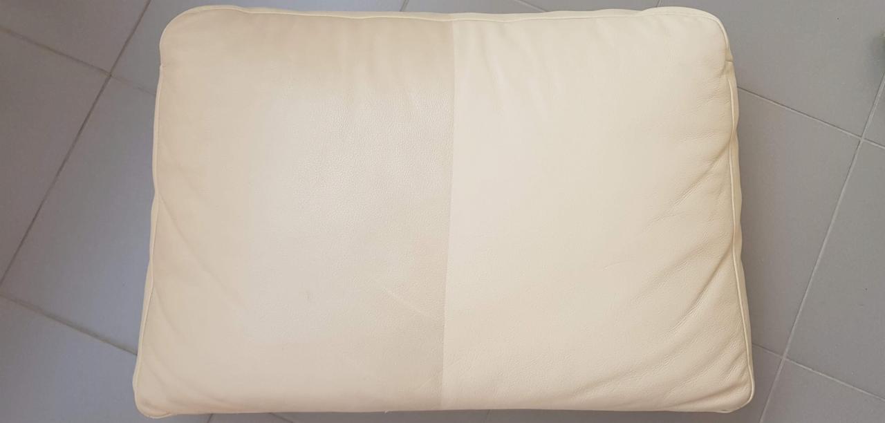 Poduszka z jasnej skóry ekologicznej, widoczna różnica w czystości między lewą, wyczyszczoną połową a prawą, jeszcze nieczyszczoną częścią. Poduszka leży na szarej podłodze z płytek.