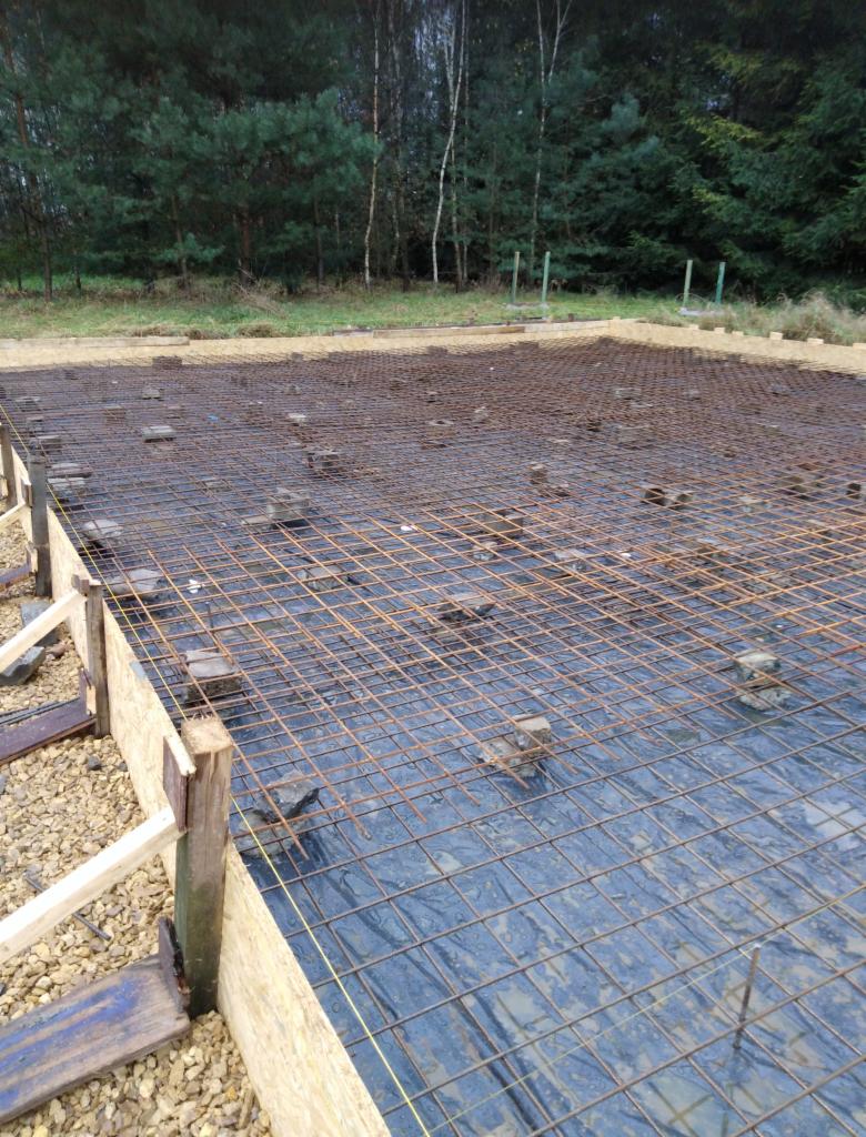 Przygotowanie zbrojenia fundamentów z deskowaniem i folią izolacyjną na działce budowlanej z widokiem na las w tle.