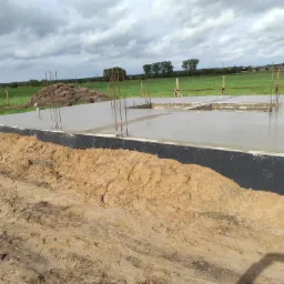 Wylany fundament z wystającymi prętami zbrojeniowymi, zabezpieczony czarną folią hydroizolacyjną, z widoczną ziemią i zielonym polem w tle pod pochmurnym niebem.