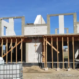 Surowy stan domu w trakcie budowy, widoczne bloczki, drewniane szalunki, rusztowanie, materiały budowlane i zbiornik na wodę na działce z widokiem na okolicę.