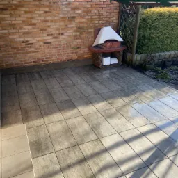 Szara kostka brukowa ułożona na tarasie przylegającym do ceglanego budynku, z widocznym piecem ogrodowym i fragmentem zielonego żywopłotu w tle, częściowo mokra powierzchnia odbija promienie...