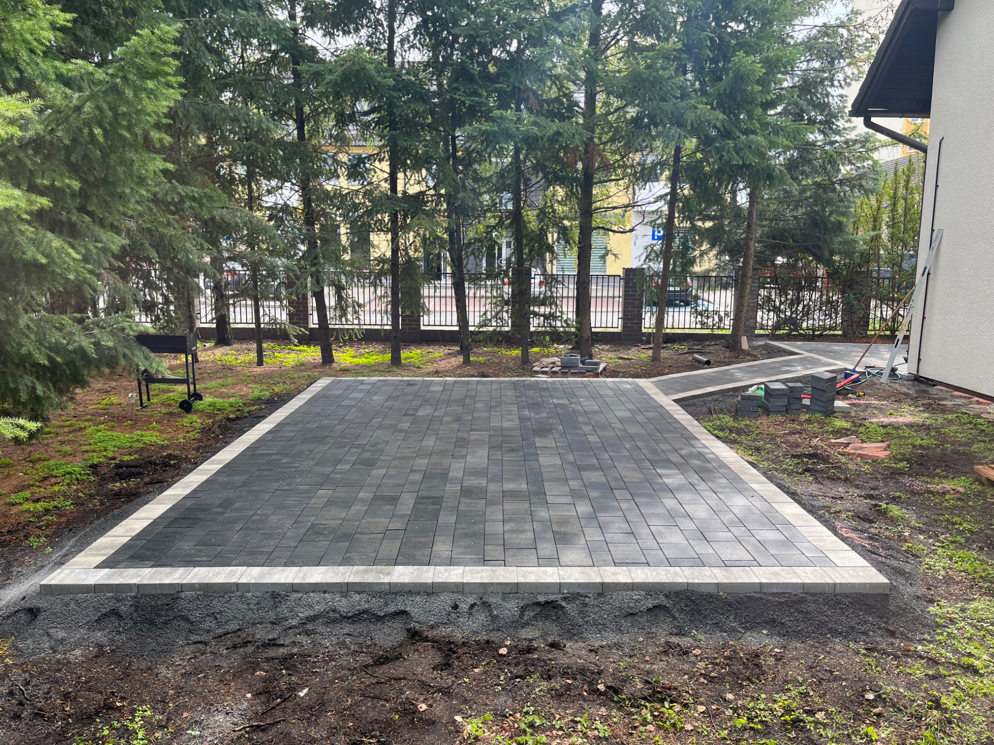 Nowo ułożona, prostokątna nawierzchnia z ciemnoszarej kostki brukowej obramowana jaśniejszymi elementami, widoczna na tle zieleni i drzew iglastych, z przygotowanym podłożem z drobnego kruszywa.