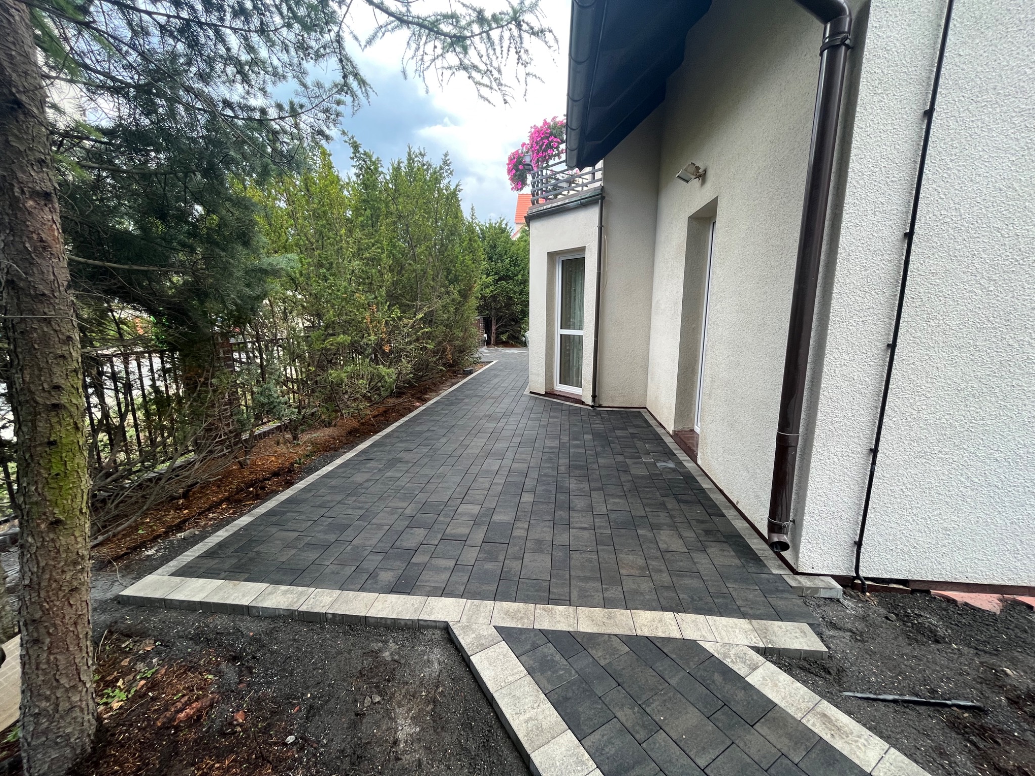 Nowo ułożona, grafitowa kostka brukowa z jasną obwódką, prowadząca wzdłuż ściany domu z balkonem ozdobionym różowymi kwiatami, widok z poziomu gruntu.
