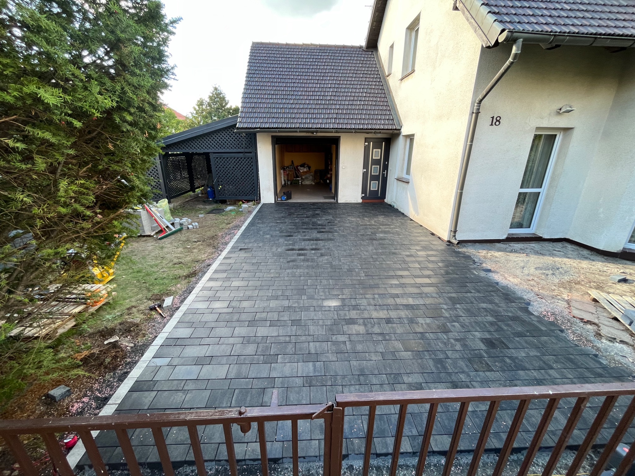 Ułożona, ciemnoszara kostka brukowa na podjeździe przed domem z numerem 18, widok zza brązowego ogrodzenia, w tle garaż i elementy ogrodu.