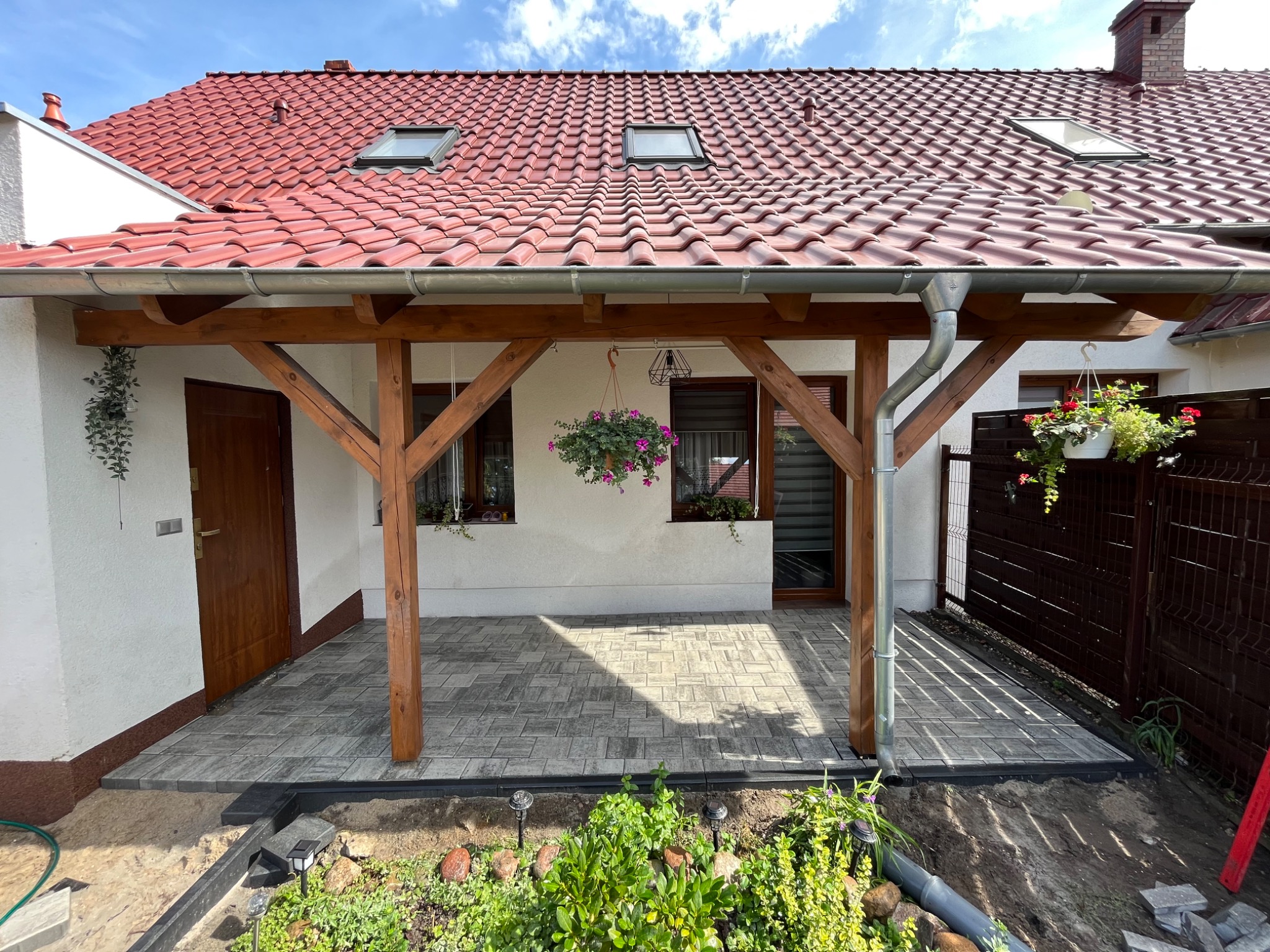 Zadaszony taras z kostki brukowej w odcieniach szarości, drewniana konstrukcja wsparta na słupach, wiszące donice z kwiatami, widok na ogród z niską roślinnością i lampami solarnymi.