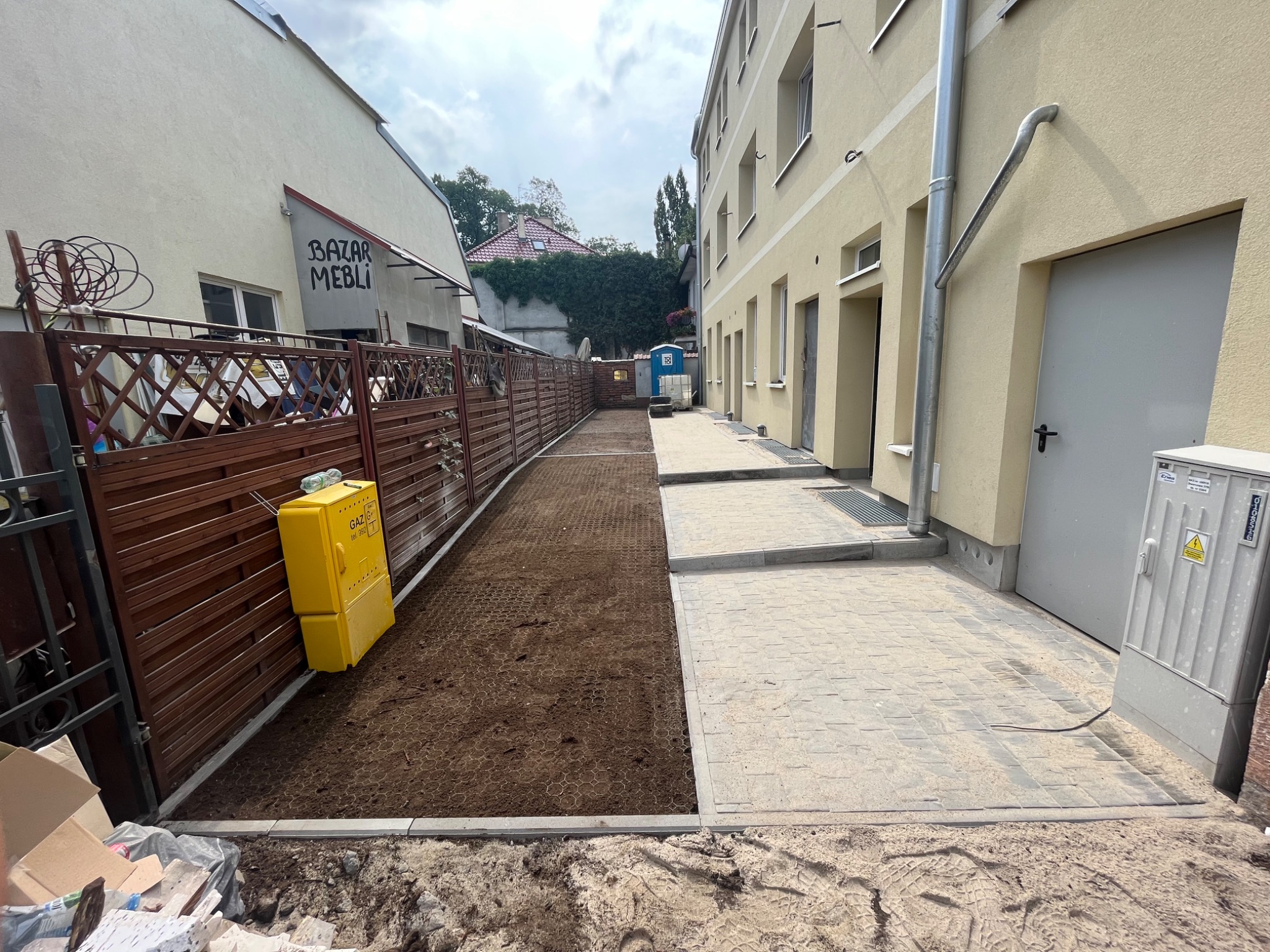 Długi, wąski dziedziniec z częściowo ułożoną szarą kostką brukową po prawej stronie i przygotowanym podłożem z geokratą wypełnioną ziemią po lewej, żółta skrzynka gazowa przy drewnianym płocie...