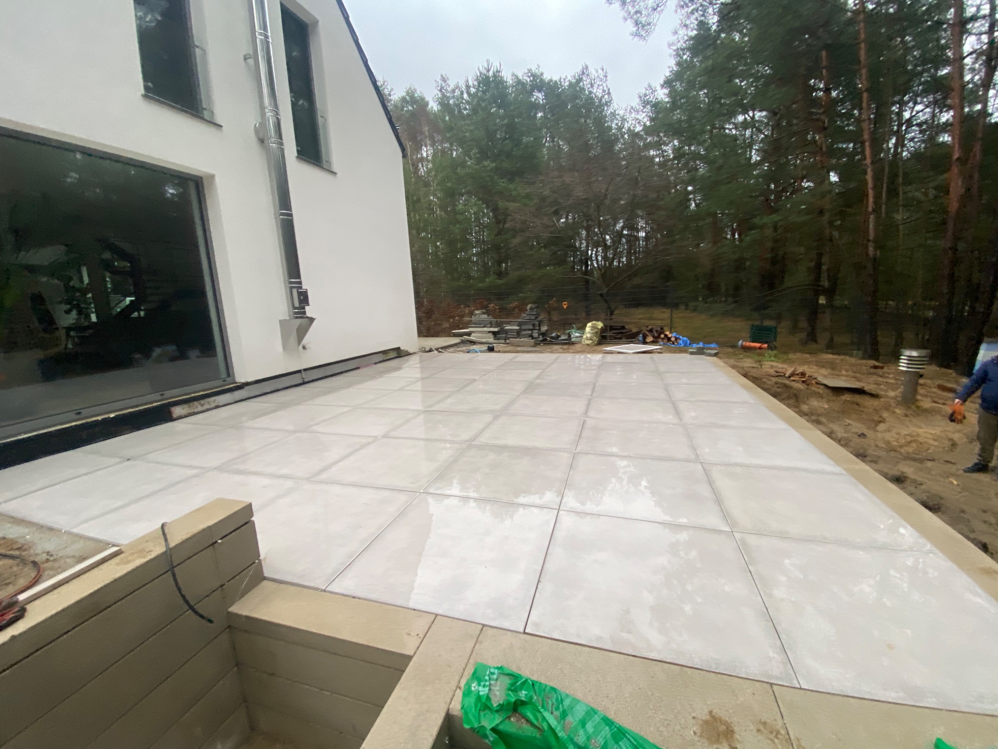Nowoczesny taras z jasnych, kwadratowych płyt, częściowo ułożonych, z widocznym przygotowaniem podłoża i elementami wykończeniowymi, na tle lasu i białego domu.