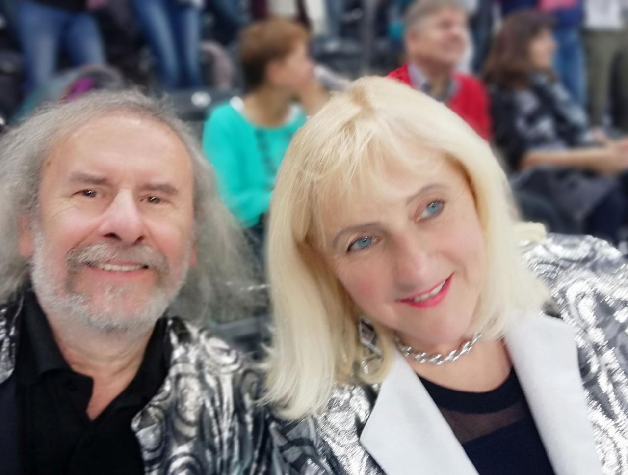 Portret dwojga uśmiechniętych artystów w srebrnych, błyszczących strojach na tle widowni.