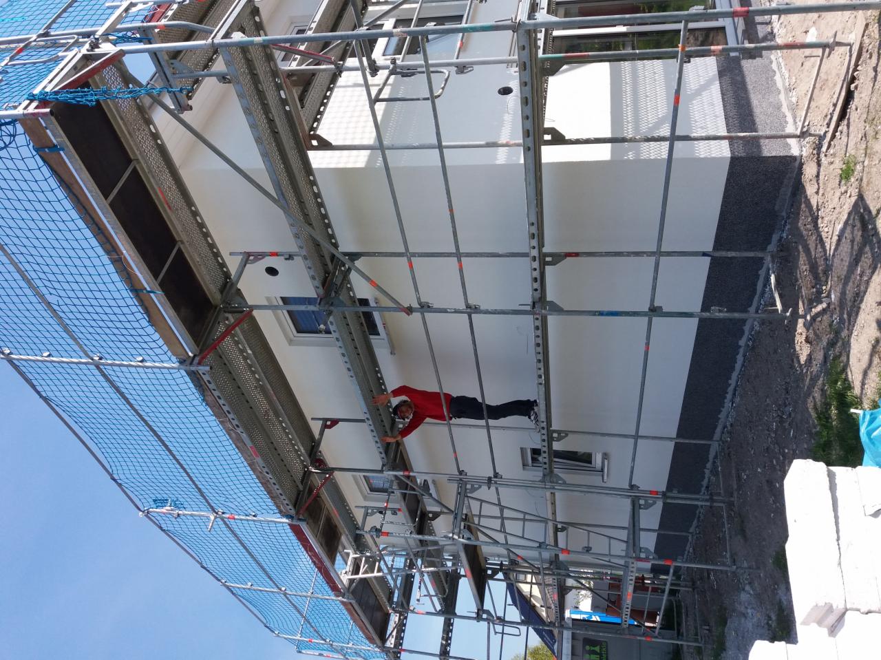 Pracownik w czerwonej bluzie na rusztowaniu podczas wykonywania prac elewacyjnych na budynku z oknami, widoczna siatka zabezpieczająca.