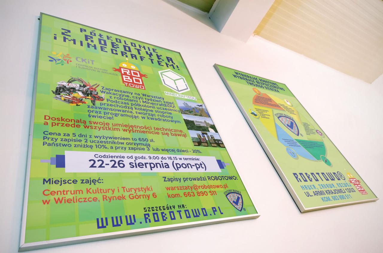 Dwa plakaty informacyjne na białej ścianie. Lewy plakat reklamuje półkolonie robotyki i Minecrafta, z robotem z klocków na grafice. Prawy plakat promuje bezpieczeństwo dziecka.