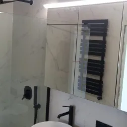 Nowoczesna łazienka z okrągłą umywalką nablatową, baterią w kolorze czarnym, grzejnikiem drabinkowym odbijającym się w lustrze i oświetleniem punktowym.