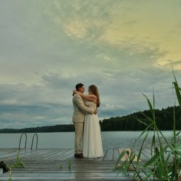 filmslubny.eu - Para w eleganckich strojach, obejmująca się na mokrym drewnianym pomoście nad jeziorem, otoczona zielonymi trzcinami. W tle zachmurzone niebo z delikatnym, złotawym światłem.