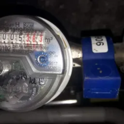 Licznik wody z niebieskim zaworem odcinającym, wskazujący zużycie 5,383 m3, zamontowany na metalowej rurze w ciemnym otoczeniu.