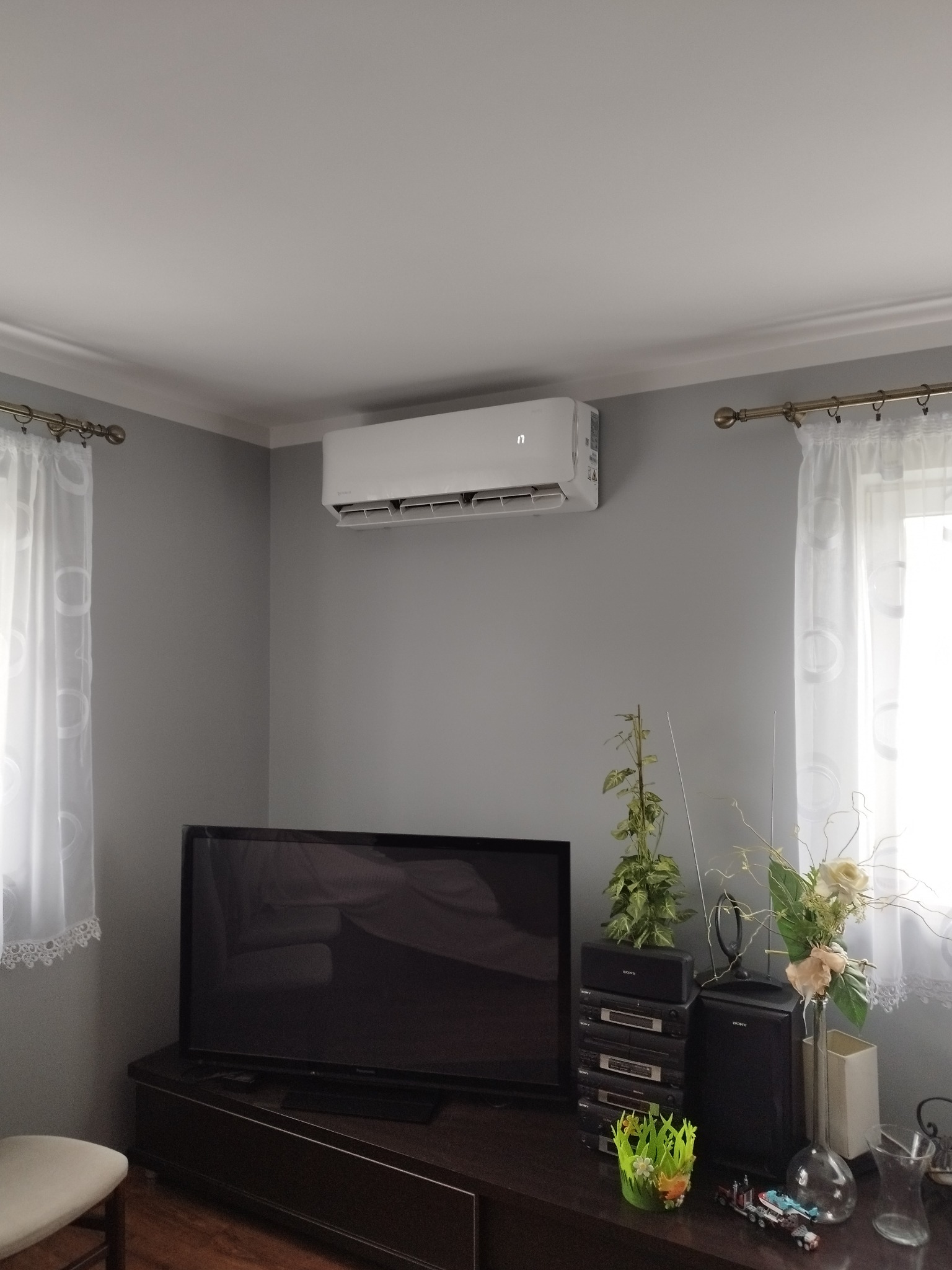 Biała klimatyzacja na szarej ścianie w pokoju z telewizorem i kwiatami. Widok z przodu, stonowane oświetlenie.