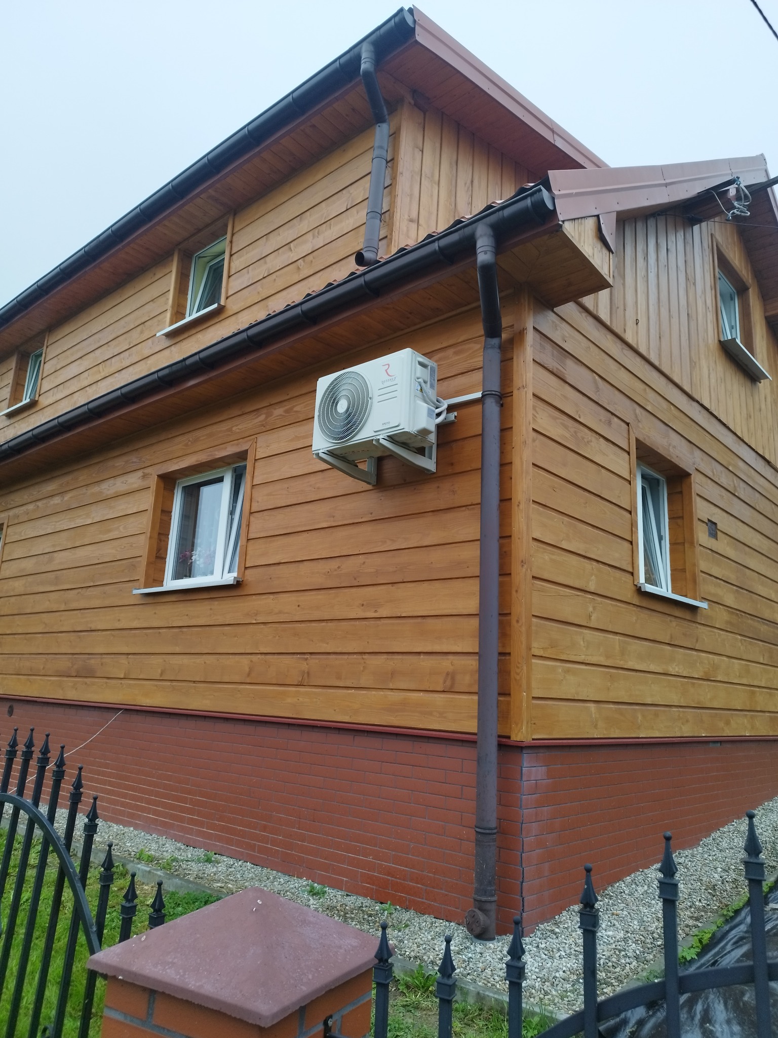 Boczny widok domu z drewnianą elewacją i zamontowaną klimatyzacją na ścianie. Widoczne okna z białymi ramami, rynna i fragment ogrodzenia.