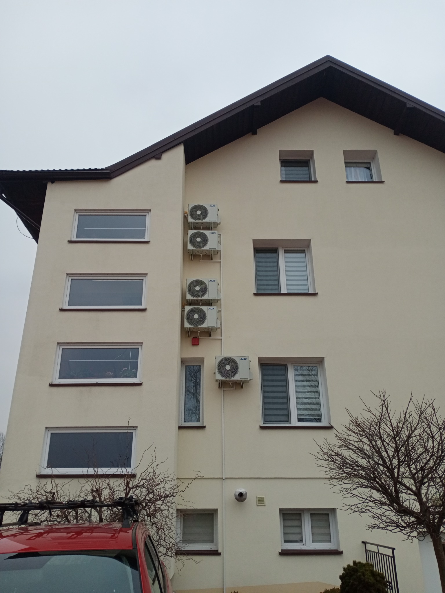 Elewacja budynku z zamontowanymi klimatyzatorami AUX, widoczne okna i fragment czerwonego samochodu. Montaż klimatyzacji w budynku wielorodzinnym.