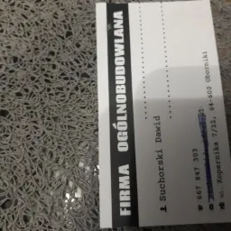 Wizytówka firmy ogólnobudowlanej Suchorski Dawid na szarej, teksturowanej powierzchni. Zawiera dane kontaktowe: numer telefonu i adres.