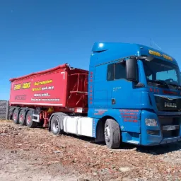 Niebiesko-czerwona ciężarówka MAN z przyczepą, na której widnieje napis 'Sprzedaż: piach, żwir, tłuczeń, ziemia...', zaparkowana na placu z gruzem, w tle betonowy płot.