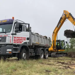 Żółta koparka JCB załadowuje ziemię do skrzyni ładunkowej szarej ciężarówki MAN firmy BUD-TRANS na tle łąki i drzew.