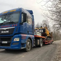 Niebieska ciężarówka z napisem BUD-TRANS Częstochowa przewozi żółtą koparkę JCB na czerwonej platformie transportowej polną drogą w pochmurny dzień.