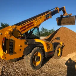 Żółta ładowarka teleskopowa JCB 540-140 z łyżką nad stertą drobnego kruszywa w słoneczny dzień, widoczny cień osoby robiącej zdjęcie.