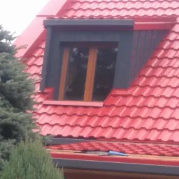 Czerwony dach z blachodachówki z lukarną o ciemnej obudowie, widoczny komin z cegły i fragment zielonego iglaka.