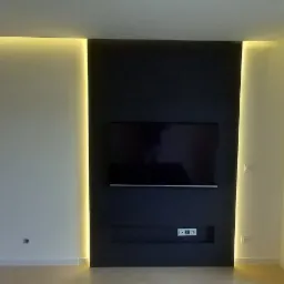 Nowoczesny salon z czarną ścianą TV podświetloną LED, widok na balkon i zabudowę kuchenną. Minimalistyczny design, jasne ściany i podłoga. Oświetlenie punktowe.