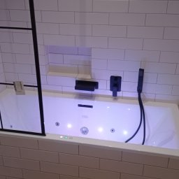 Filip Janowski - Biała wanna z hydromasażem, obudowana białymi, prostokątnymi płytkami, z czarną armaturą i szklanym parawanem z czarną ramą, oświetlona niebieskim światłem.