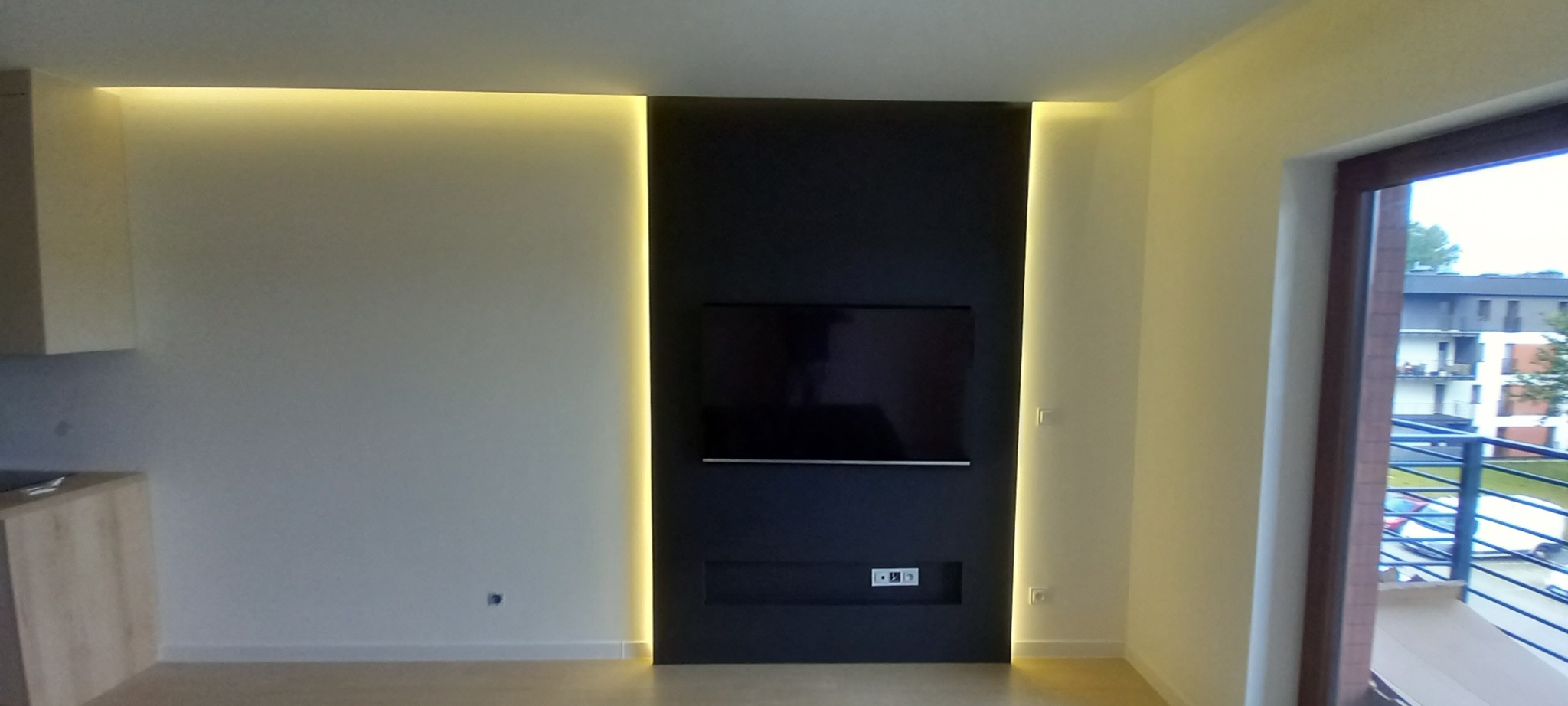 Nowoczesny salon z czarną ścianą TV podświetloną LED, widok na balkon i zabudowę kuchenną. Minimalistyczny design, jasne ściany i podłoga. Oświetlenie punktowe.