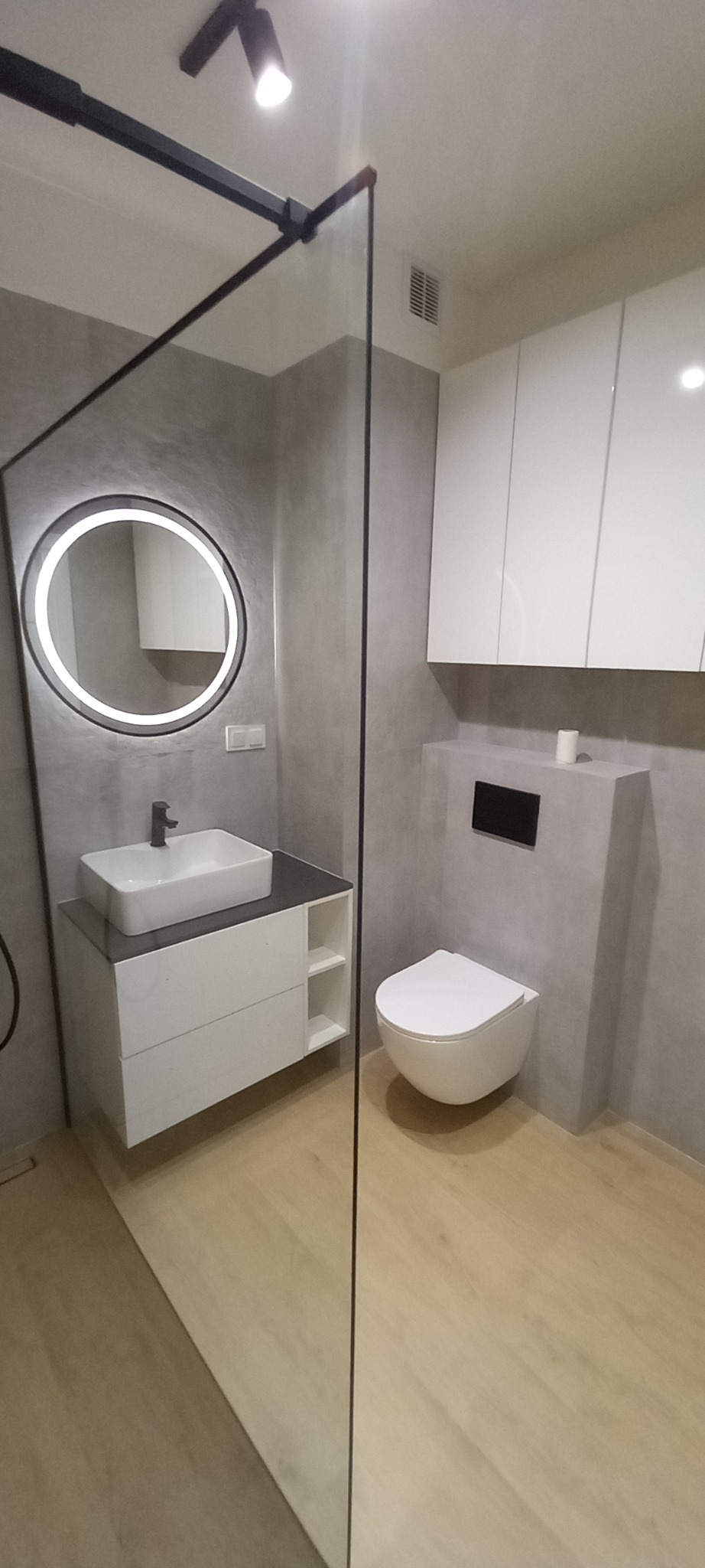 Nowoczesna łazienka w szarościach z okrągłym lustrem LED, wiszącą toaletą i drewnopodobną podłogą. Kabina prysznicowa typu walk-in z czarną ramą.