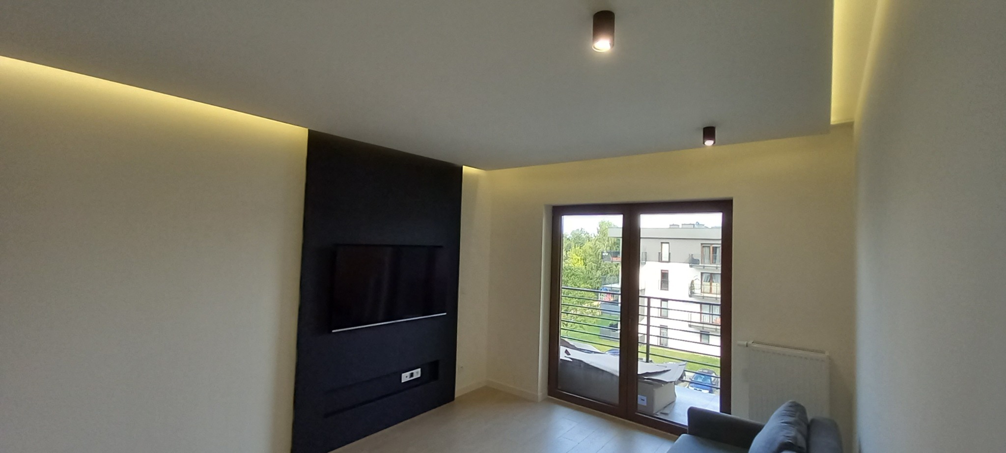 Nowoczesny salon z czarną ścianą TV, subtelnym oświetleniem LED i wyjściem na balkon. Minimalistyczny design, jasne ściany, podłoga i punktowe lampy.