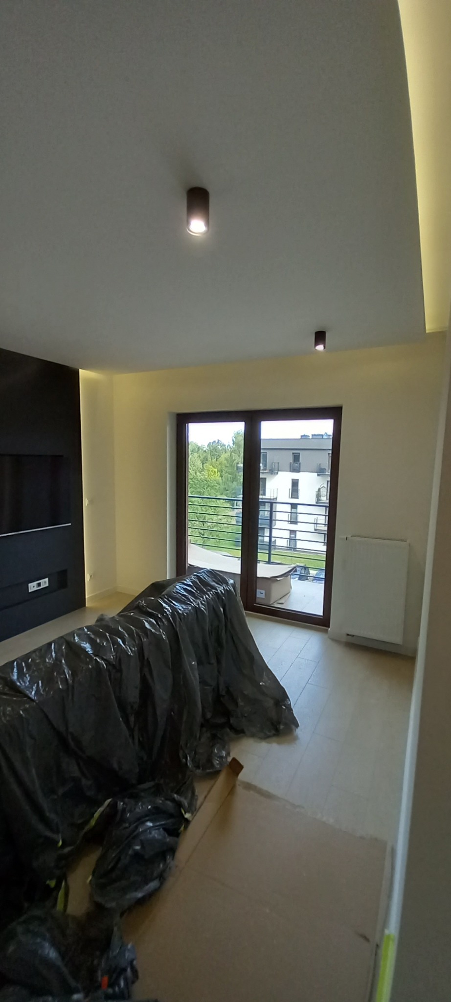 Wnętrze mieszkania w trakcie prac wykończeniowych. Sofa zabezpieczona folią, widoczne kartony, okno balkonowe z widokiem na zieleń i budynek.