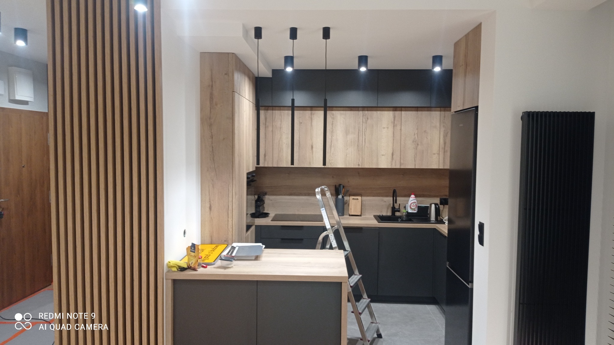 Nowoczesna kuchnia z drewnianymi szafkami i czarnymi akcentami, widoczna drabina malarska sugeruje prace wykończeniowe, a drewniane lamele oddzielają przestrzeń.