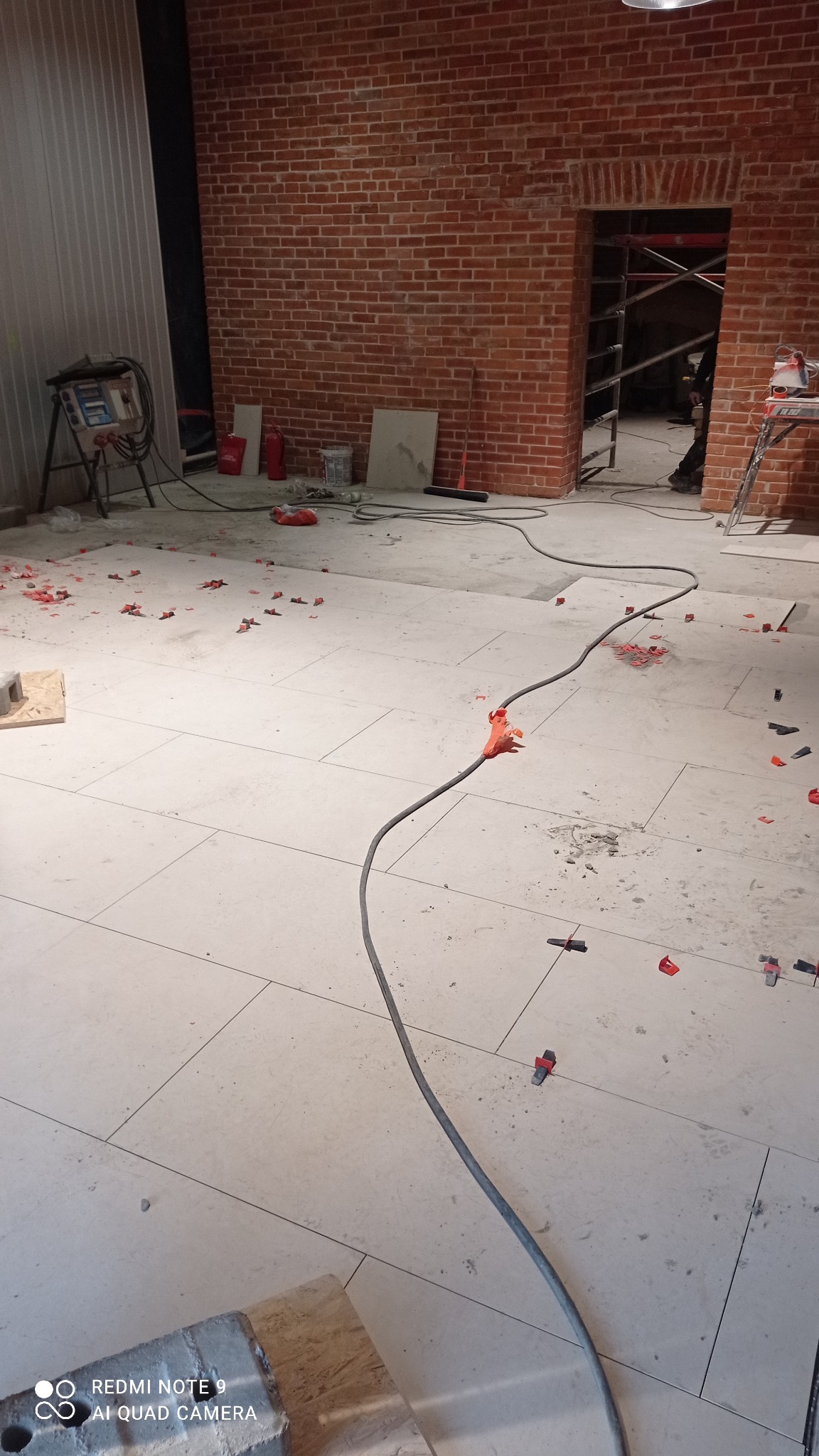 Wnętrze w trakcie prac wykończeniowych: ułożona jasna podłoga z płytek, fragment ściany z czerwonej cegły, widoczne narzędzia i akcesoria glazurnicze oraz przewód elektryczny. W tle rusztowanie...