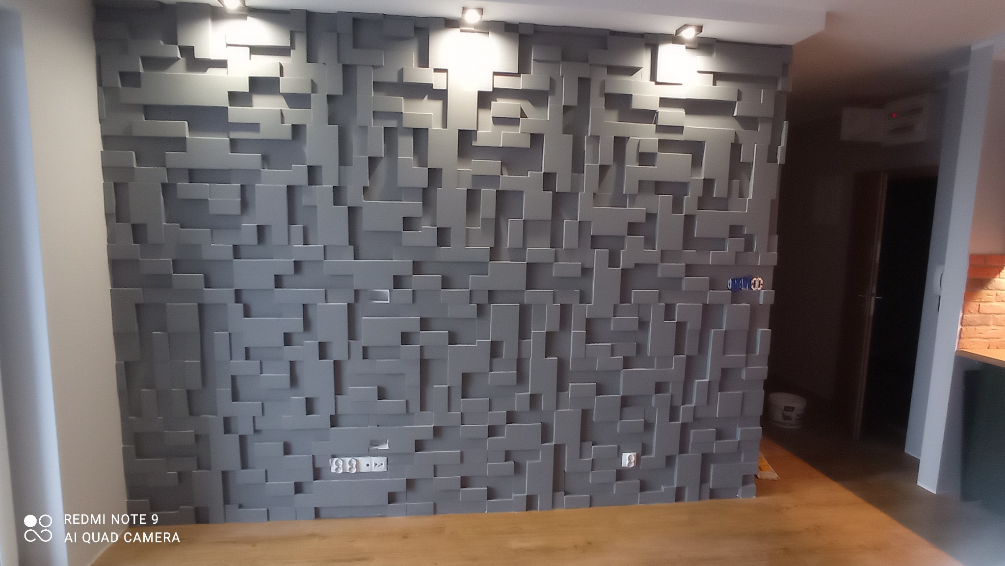 Szara ściana dekoracyjna z trójwymiarowych paneli w kształcie prostopadłościanów, podświetlona punktowymi lampami sufitowymi, z widocznymi gniazdkami elektrycznymi i fragmentem podłogi z paneli.