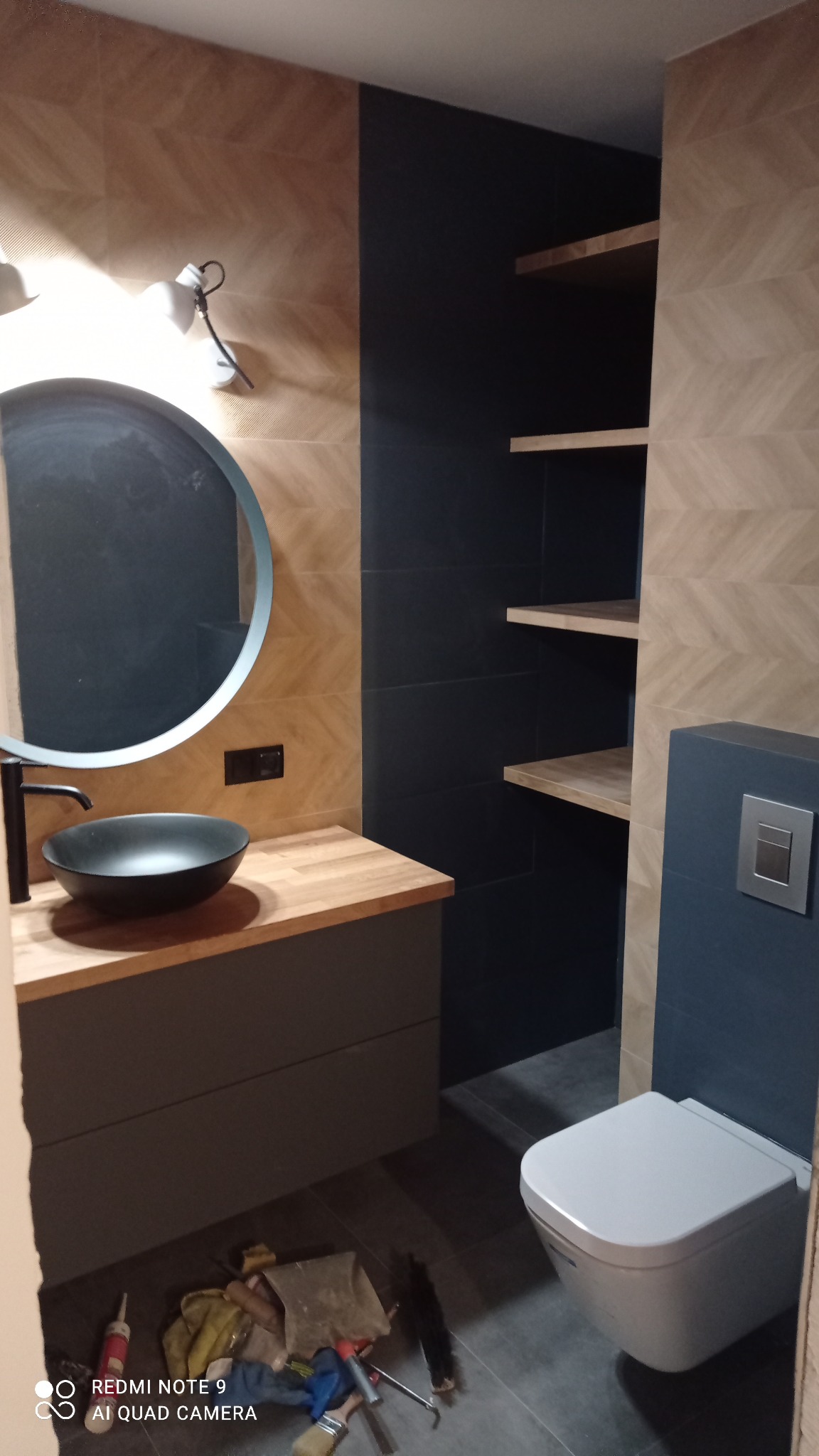 Nowoczesna łazienka z okrągłym lustrem w niebieskiej ramie, czarną umywalką nablatową i drewnianym blatem, płytki imitujące drewno na ścianach, czarne płytki we wnęce z drewnianymi półkami, wisząca...