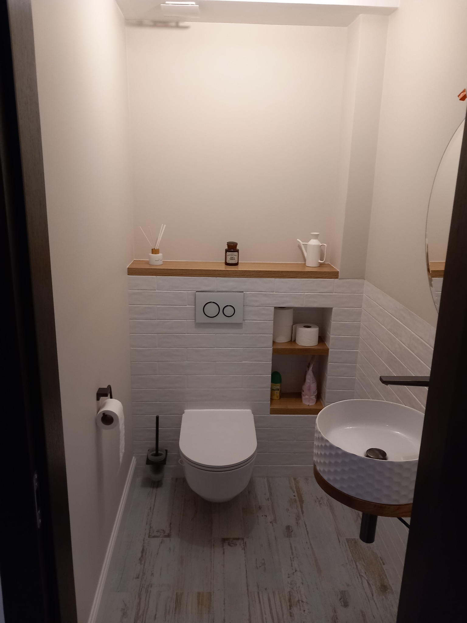 Nowoczesna łazienka z wiszącą miską WC, umywalką nablatową o fakturze plastra miodu, drewnianymi akcentami i półkami w ścianie wyłożonej białymi, poziomymi płytkami.