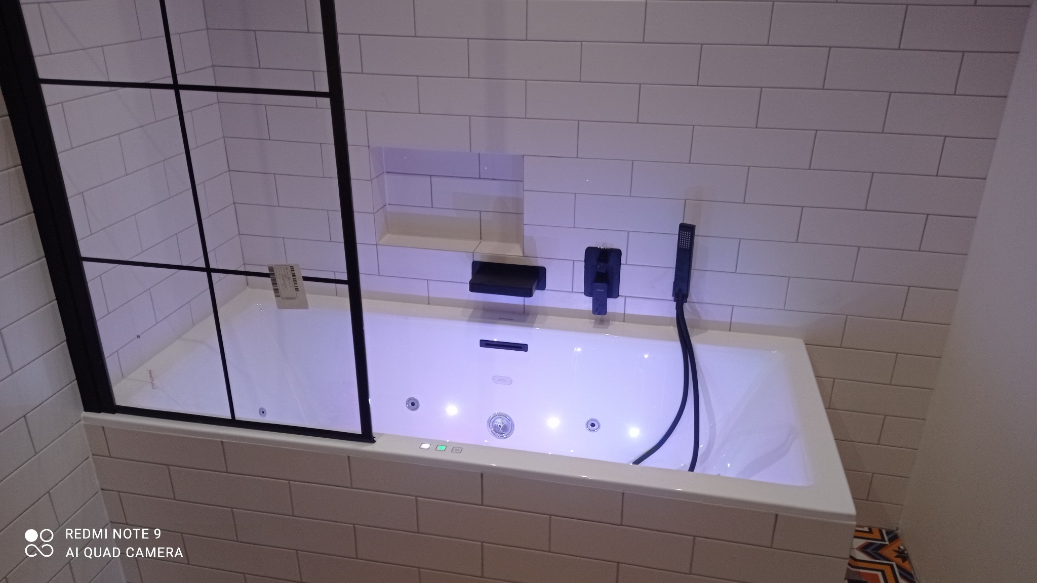 Biała wanna z hydromasażem, obudowana białymi, prostokątnymi płytkami, z czarną armaturą i szklanym parawanem z czarną ramą, oświetlona niebieskim światłem.