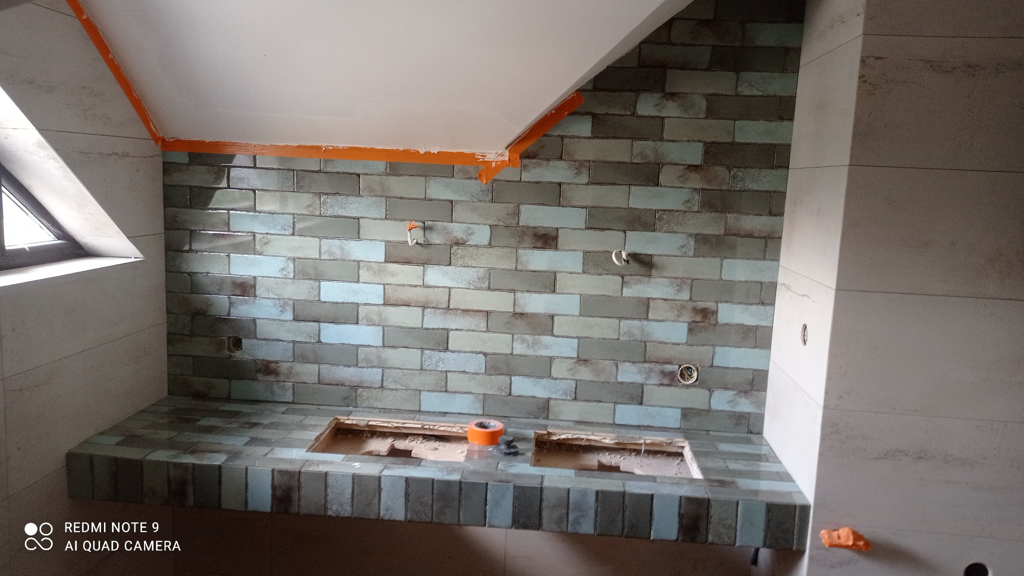 Ściana łazienki w trakcie układania płytek w odcieniach szarości i błękitu, z widocznymi otworami na instalacje i taśmą malarską na suficie, przygotowywana pod montaż umywalki.