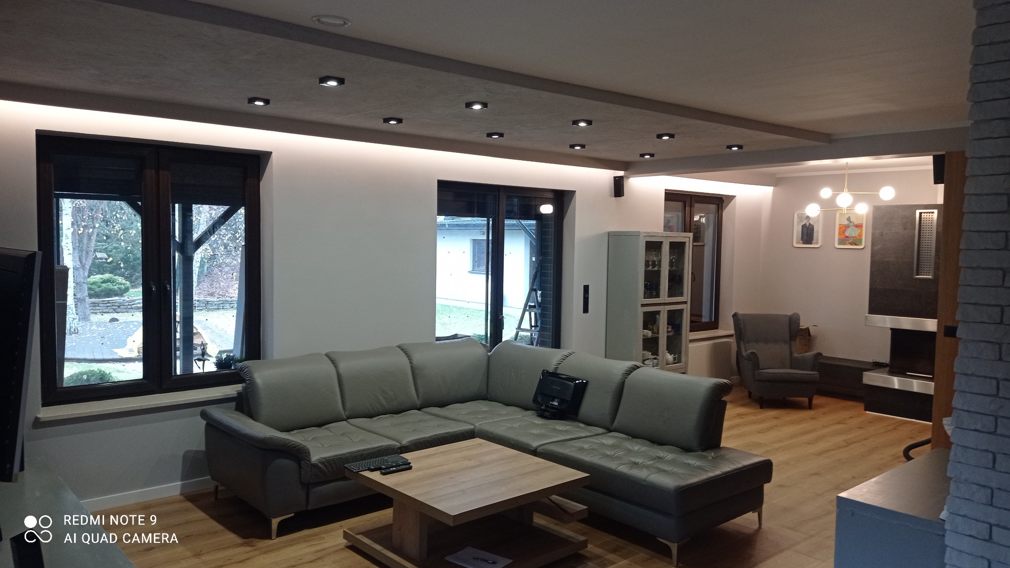 Nowoczesny salon z szarą narożną sofą, drewnianym stolikiem kawowym i oknami z ciemnymi ramami. Na suficie oświetlenie punktowe i listwa LED. Widoczny fragment kominka i fotel w tle.