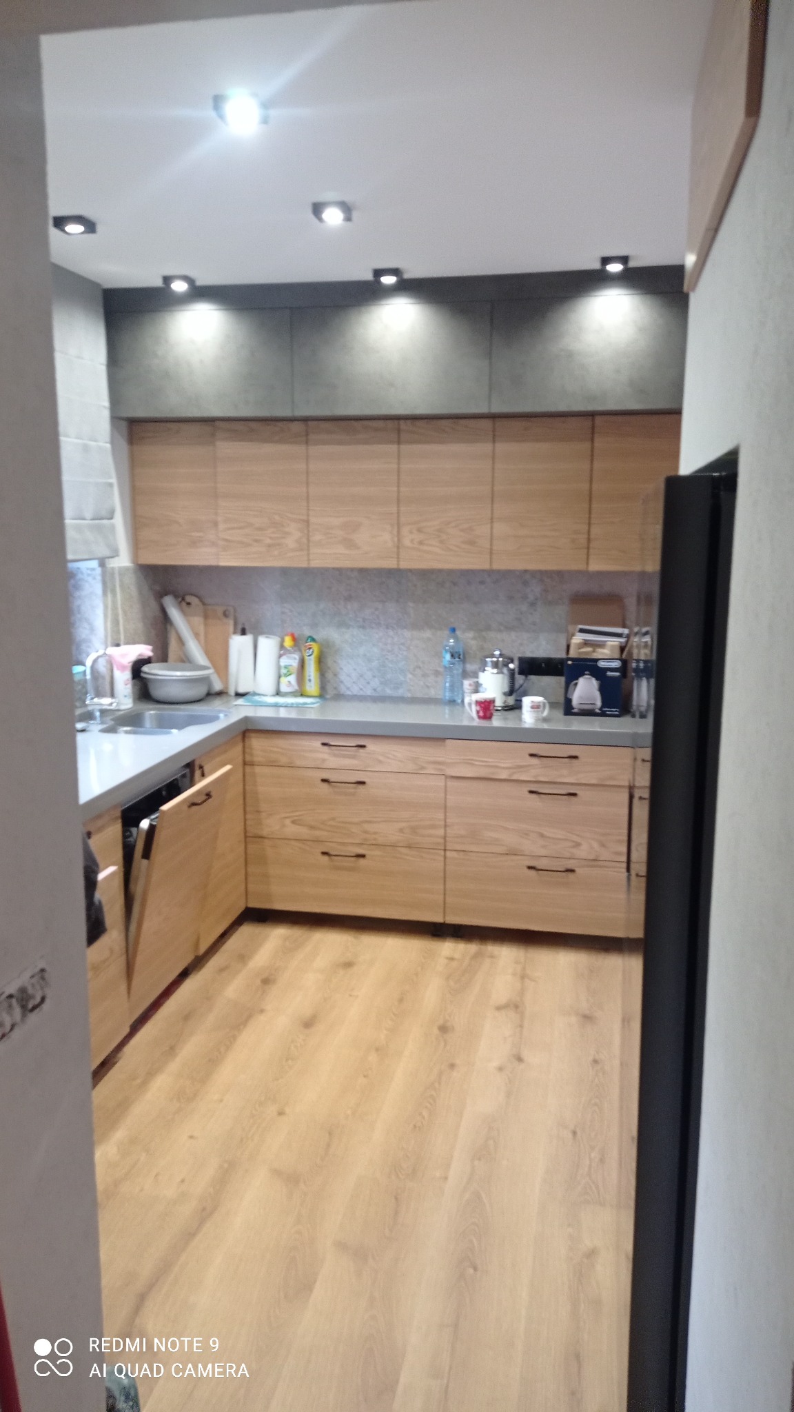 Jasna kuchnia z drewnianymi frontami szafek i szarym blatem, widoczne oświetlenie punktowe, lekko uchylona zmywarka i akcesoria kuchenne na blacie.