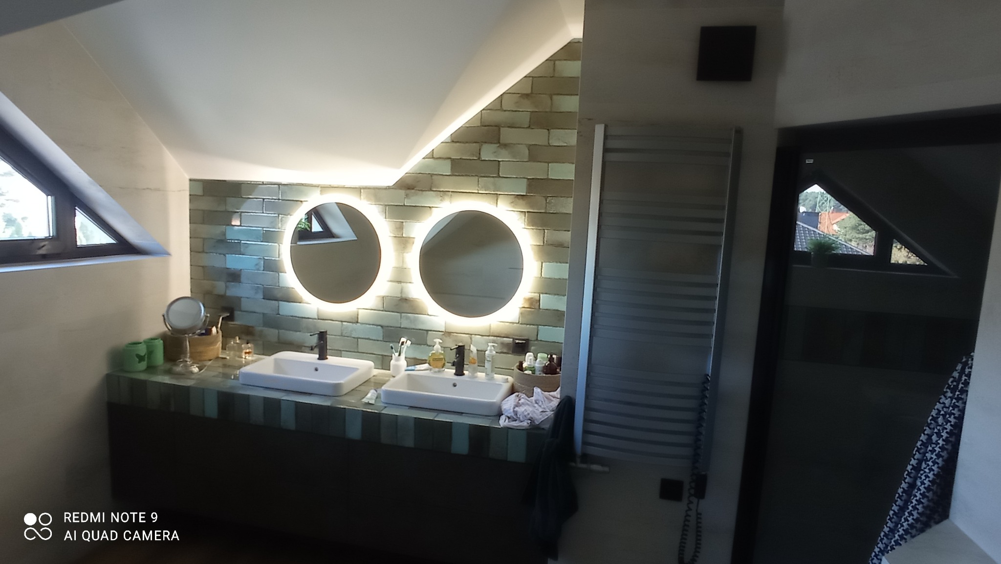 Nowoczesna łazienka na poddaszu z dwoma umywalkami, okrągłymi lustrami z podświetleniem LED, ścianą wyłożoną cegiełkami i grzejnikiem drabinkowym.